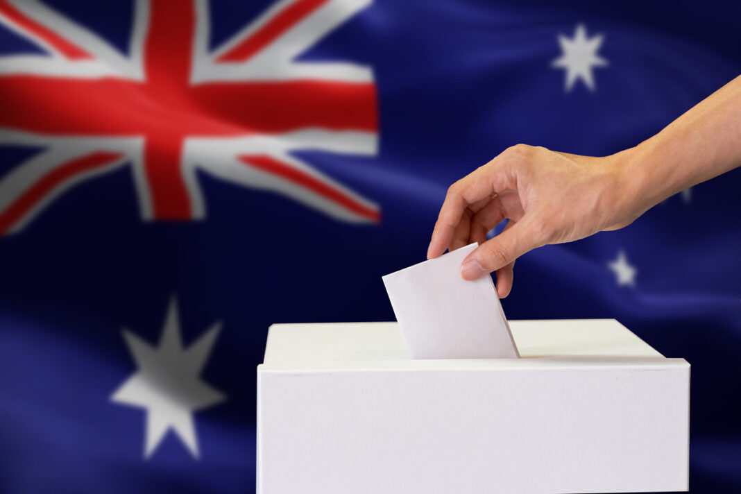 Close-up of human hand casting and inserting a vote and choosing and making a decision what he wants in polling box with Australia flag blended in background.