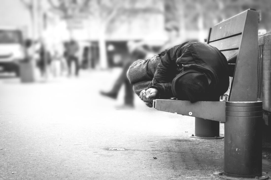Poor homeless man or refugee sleeping on the wooden bench on the urban street in the city, social documentary concept