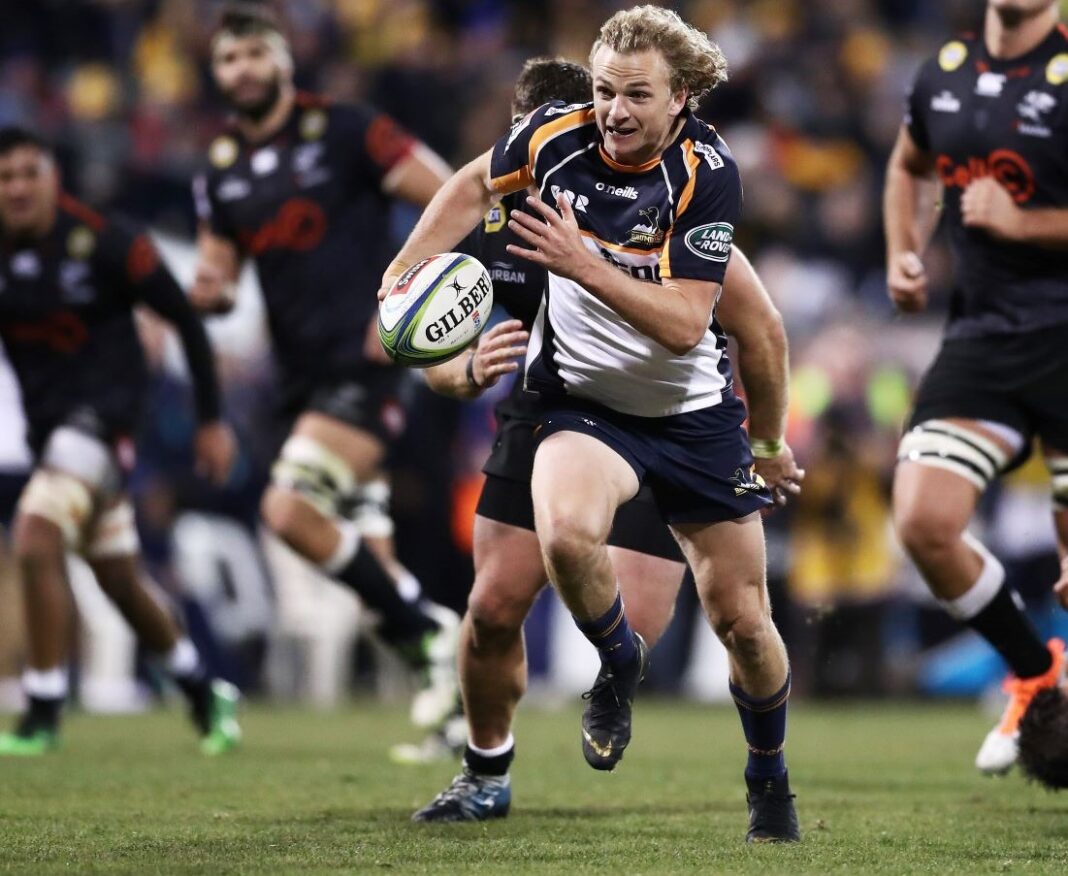 Brumbies rugby union player Joe Powell running down the field with ball in hand, blonde hair flying in the wind