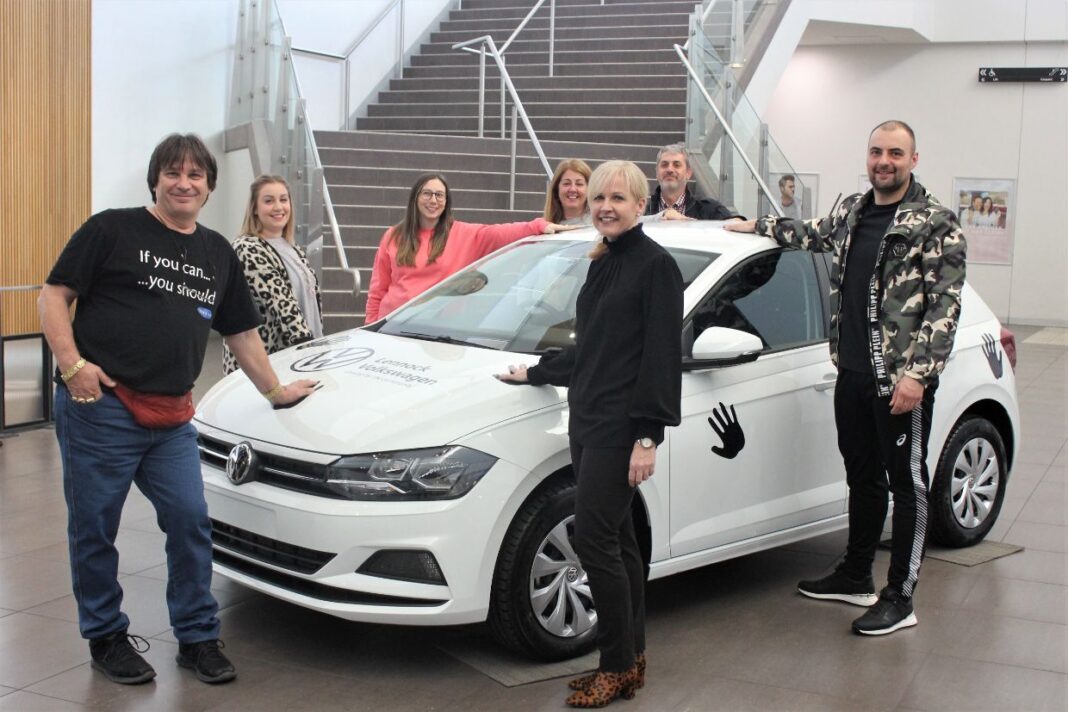 Four women and three men standing around a white Volkswagen Golf hatchback and each placing a hand on the car