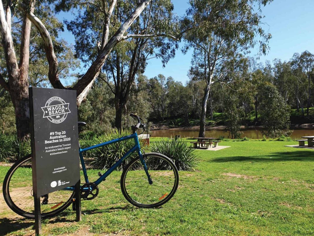 Blue bike resting against Wagga Beach sign with grass, trees and river in the background