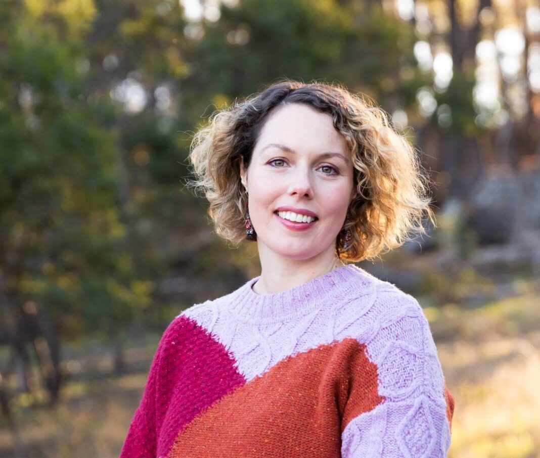 smiling woman with golden brown curly hair wearing a colourful jumper and standing in nature