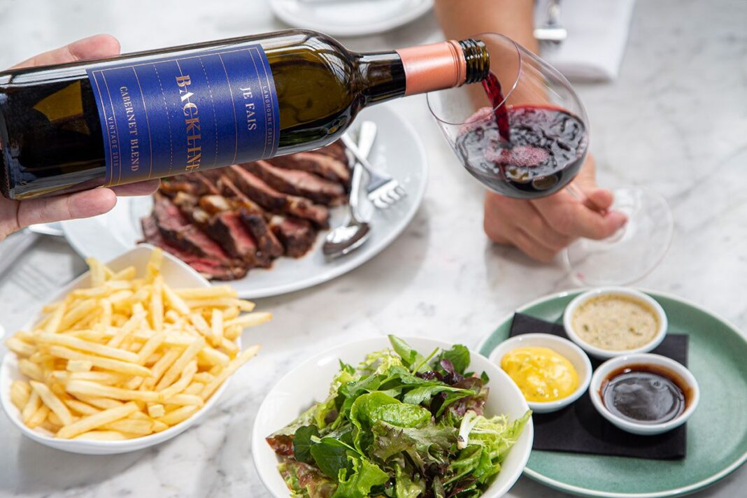 Bottle of red wine being poured into glass on table laden with meat, hot chips, salad and dips.