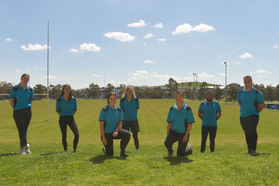 Seven athletes in blue shirts and black track pants, standing or kneeling on a sporting field.