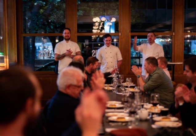 Three chefs standing at the end of a row of tables of diners in restaurant