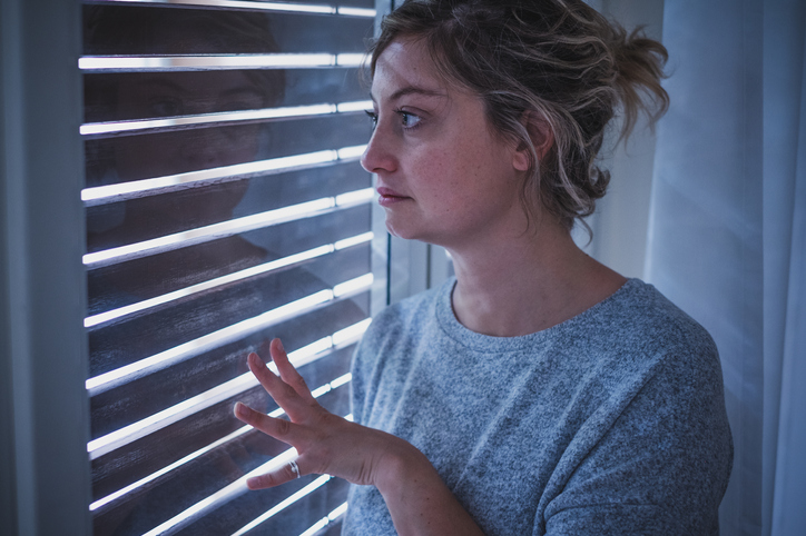 Worried young blond woman looking pensively through partially open blinds on window