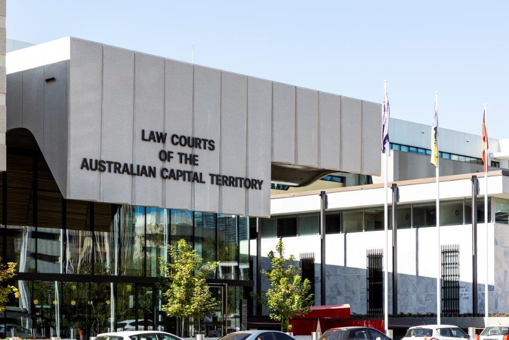 Exterior of ACT law courts building, a modern white building with lots of glass and a big black metal fence, with cars in carpark in the foreground