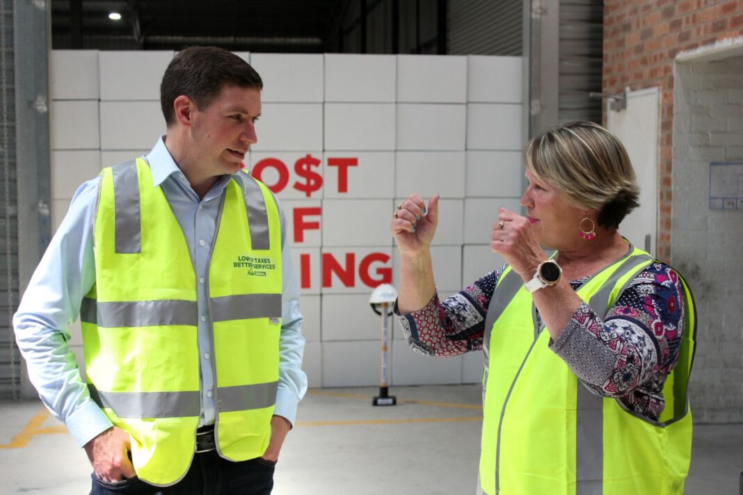 Young man and middle-aged woman wearing green hi-viz vests in front of a wall of white cardboard boxes with CO$T OF LIVING written in big red letter across the boxes