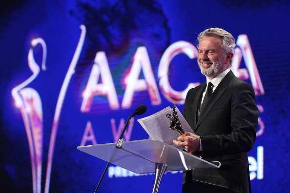 Actor Sam Neill, with wavy grey hair and a neat beard, dressed in black suit standing on stage accepting an award