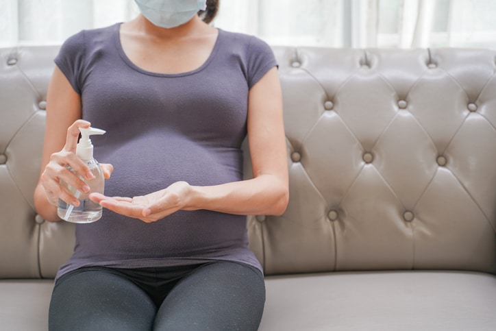 A pregnant woman wears a surgical mask is washing hands by alcohol gel on the sofa.