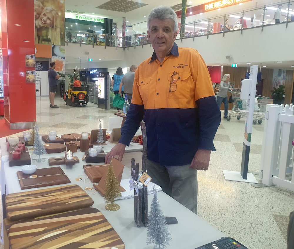 Mature man in hi-viz shirt standing near a table of woodwork items in a shopping centre