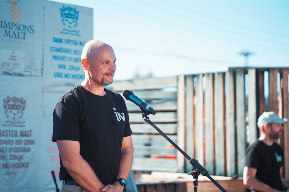 middle aged bald man in black t-shirt speaking into microphone on an outdoor stage