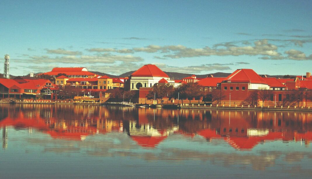 a stretch of low-rise buildings with red rooves reflected in a lake, Lake Tuggeranong in Canberra