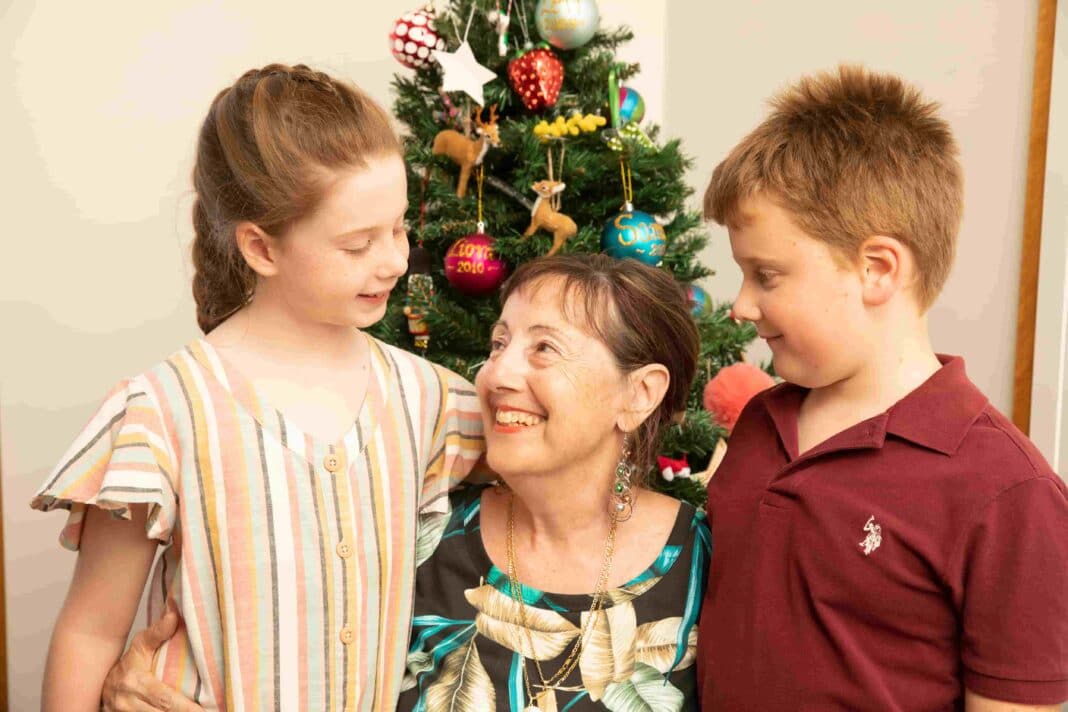 smiling grandmother embracing nine-year-old granddaughter and seven-year-old grandson in front of a Christmas tree