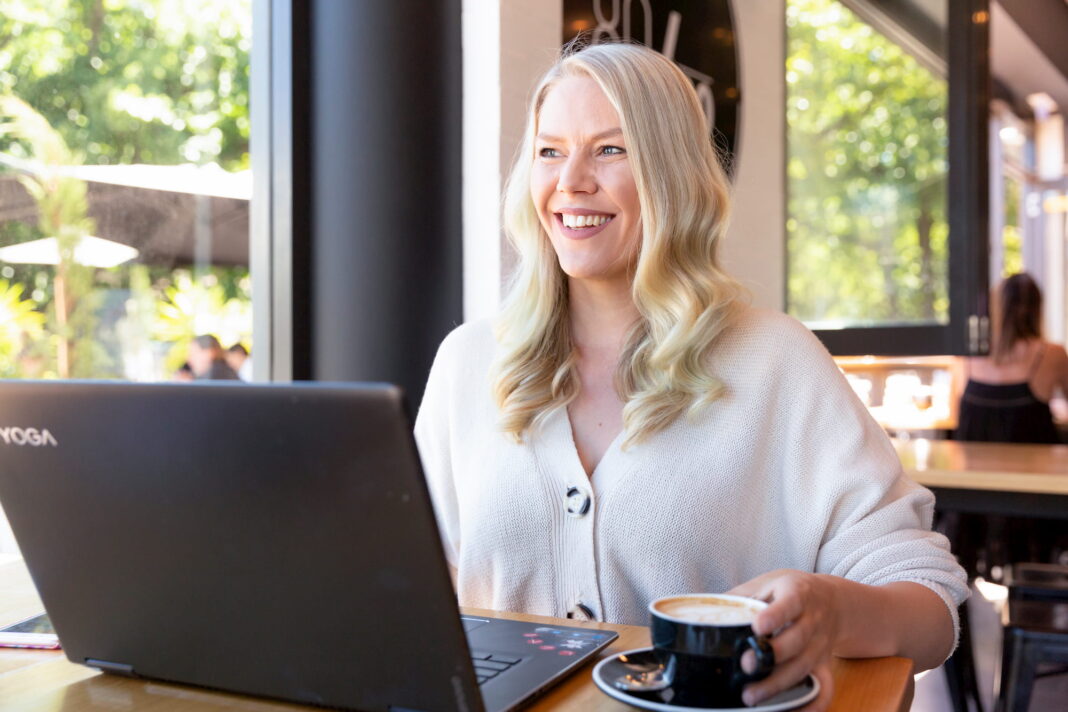 The 5:30am Club Canberra founder Emily Davidson sits at a café table smiling, with her laptop open