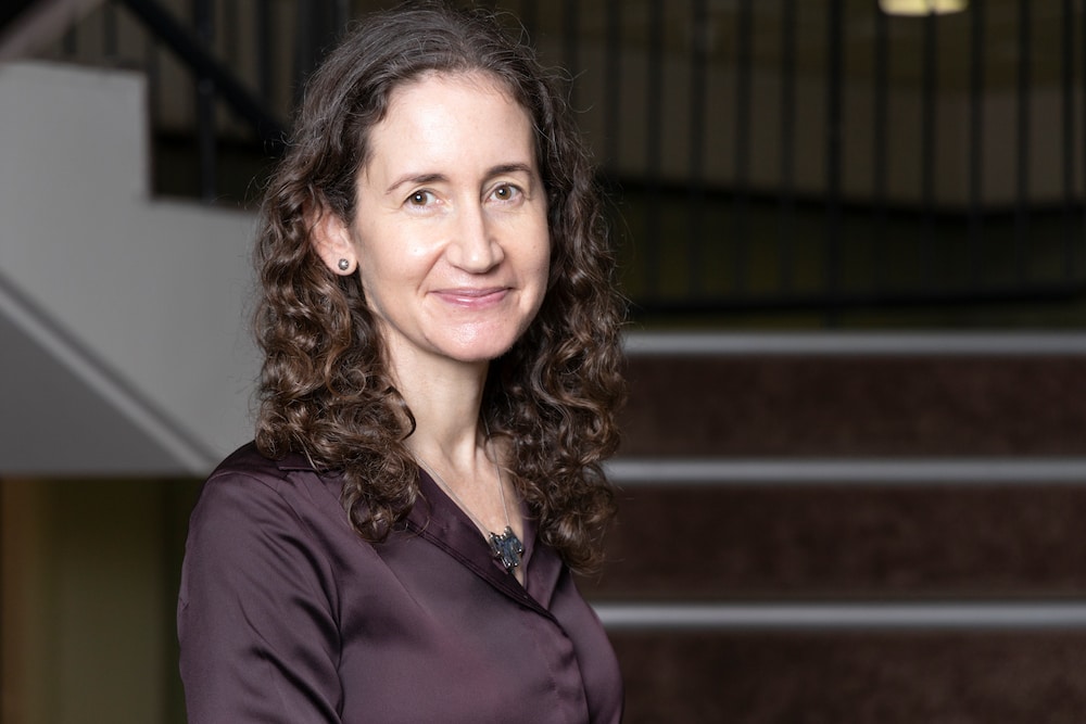 UN Women Australia's Janelle Weissman has white skin and dark brown curly hair, she wears a brown silk shirt and stands in front of a staircase
