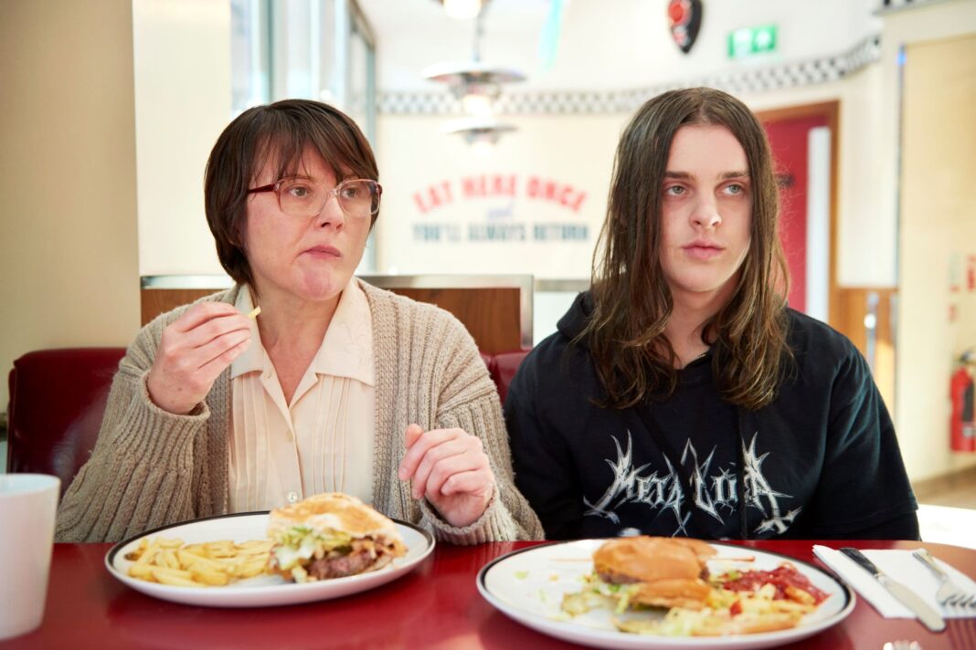 Middle aged woman in glasses and beige cardigan with a long-haired teenage boy in black eating burgers and chips at an English cafe