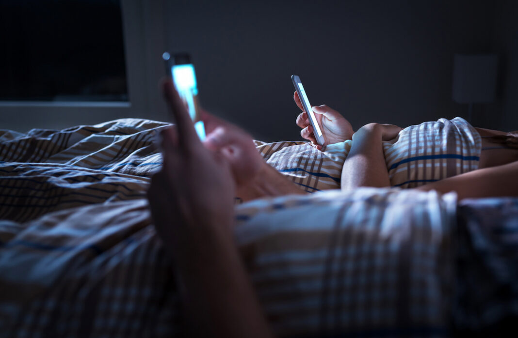 A couple lie in bed in the dark, each scrolling on their phones which provide the only light in the room. MoodOff Day challenges people worldwide to ditch this kind of behaviour.