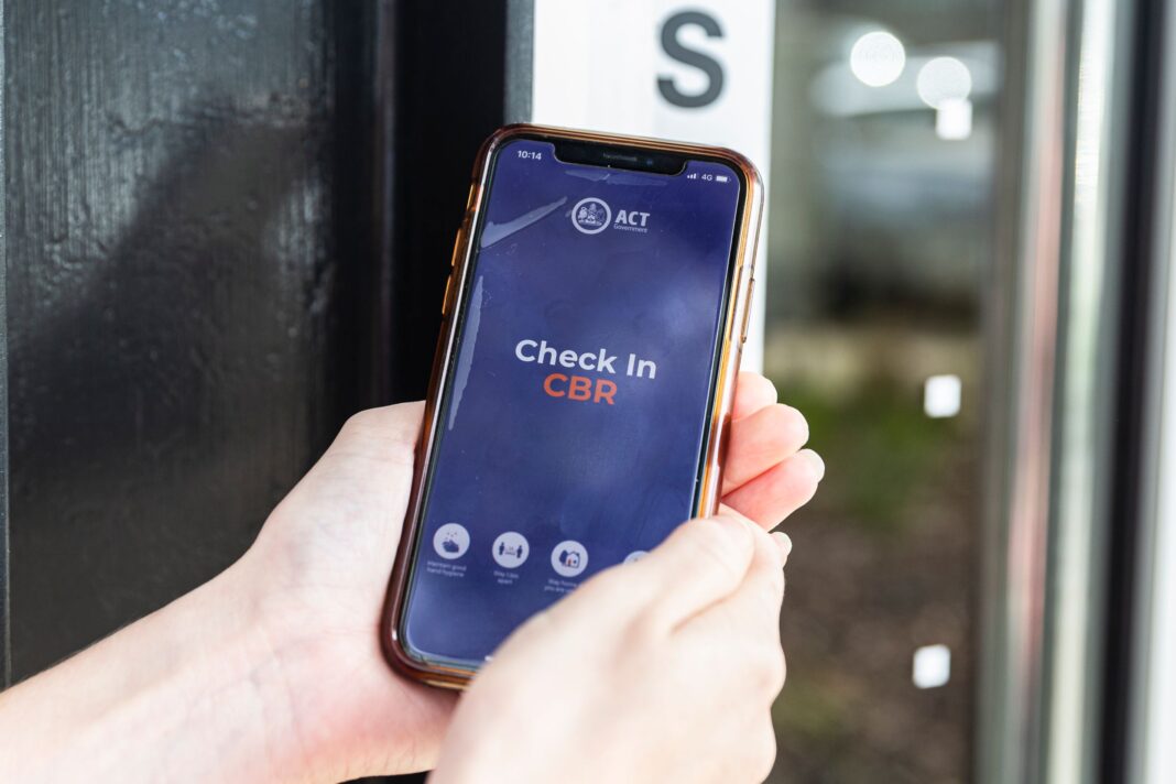 An iPhone XS displays the Check In CBR app in front of a glass door