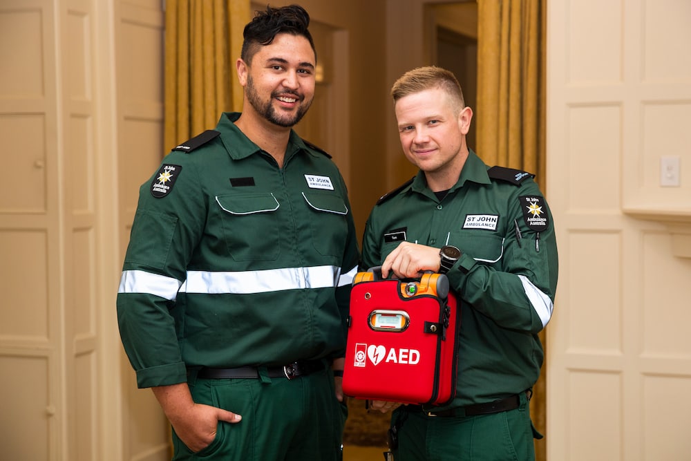 St John Ambulance volunteers in green uniforms hold a red AED (defibrillator) and smile at the camera