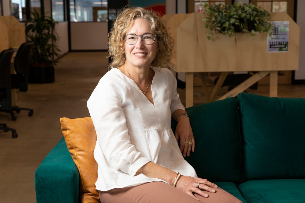 Deaf ACT secretary Louise Irvine has curly blonde hair and a white top, she sits on a green couch and smiles at the camera. She chooses to wear her cochlear implant under her hair.