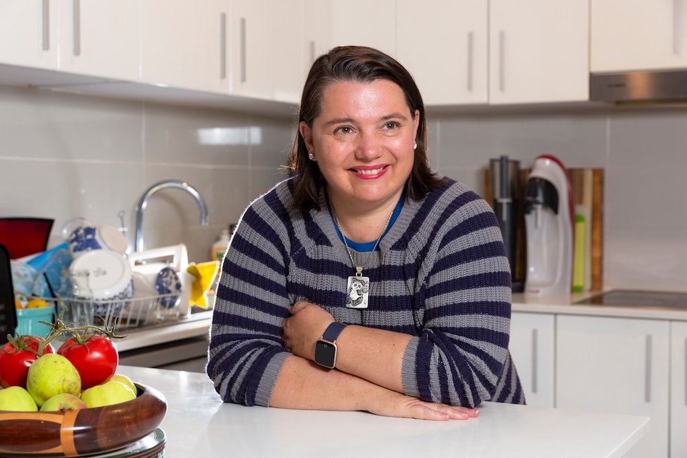YWCA Rentwell landlord Serina Bird leans on her white kitchen bench with a fruit bowl in the foreground, smiling and looking off camera, wearing a grey and black striped jumper. She has short brown hair and white skin.
