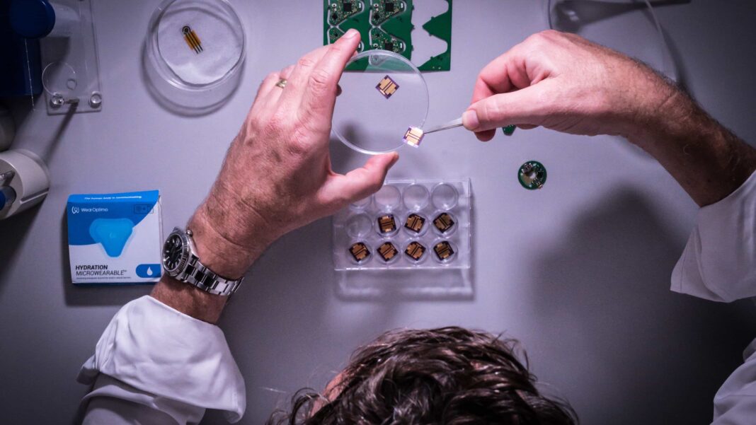 Overhead view of male scientist's hands using tweezers to place small wearable medical technology device onto a tray