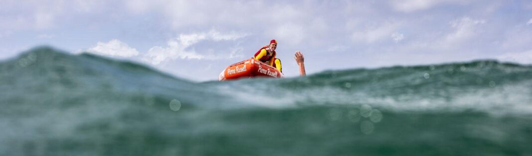 surf life saver in rescue boat approaching swimmer with one arm up in the air in ocean