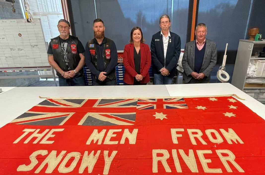 Two bikies, a female politician in a red jacket and two RSL men in suit jackets stand behind a red white an blue banner with 'The Men From Snowy River' on it