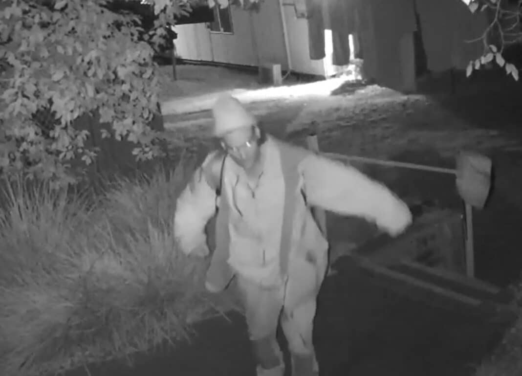 black and white CCTV footage of a man wearing a jacket, long pants, a beanie and glasses walking up a path