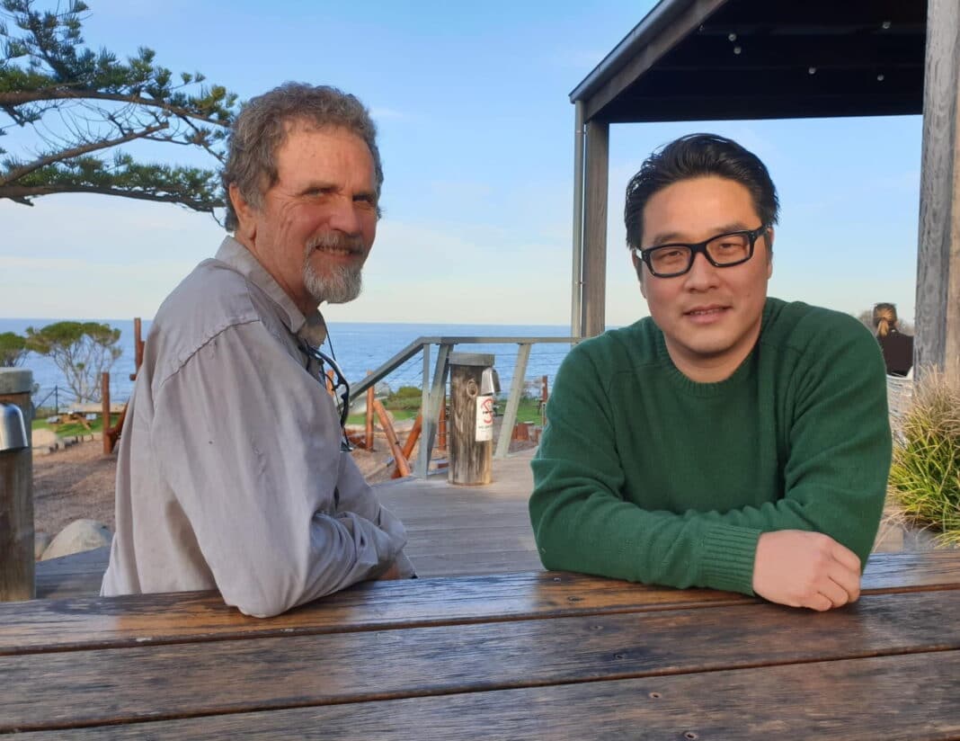 two smiling men sitting at an outdoor table overlooking the ocean at Tathra