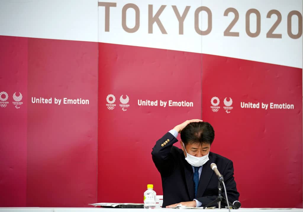 Japanese man dressed in business suit and wearing face mask scratching his head underneath a Tokyo Olympics sign