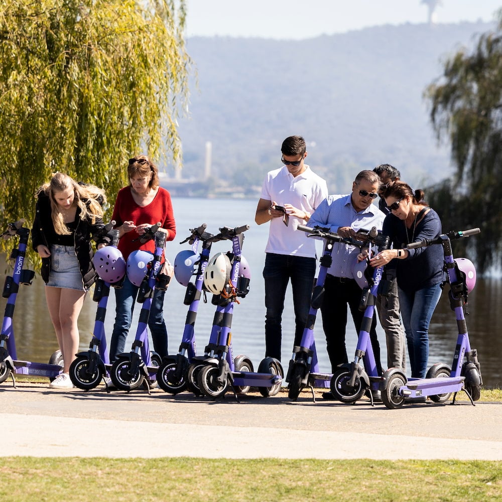 A line of several people about to hop on purple e-scooters beside Lake Burley Griffin in Canberra