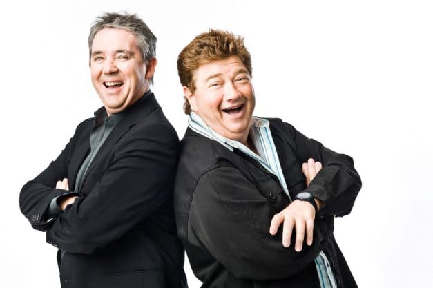 publicity shot of 2 middle aged male radio presenters standing back to back and smiling