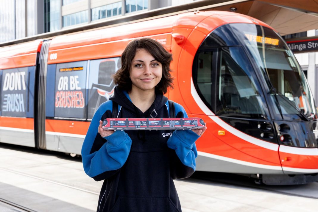Year 12 Gungahlin College Student Ada Miller holding a model of her light rail vehicle wrap design and standing in front of an actual light rail vehicle