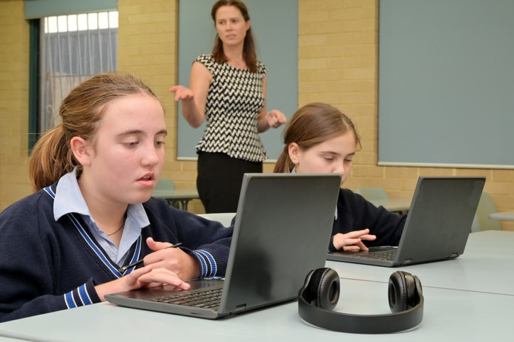 Technology teacher (Female age 30-40) teaching schoolgirls how to use laptops in school classroom.
