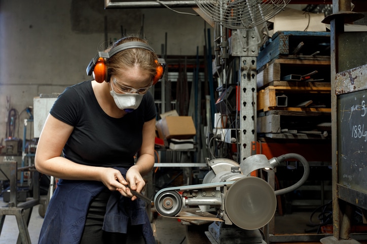 Young Australian female tradesperson using belt sander in metal workshop
