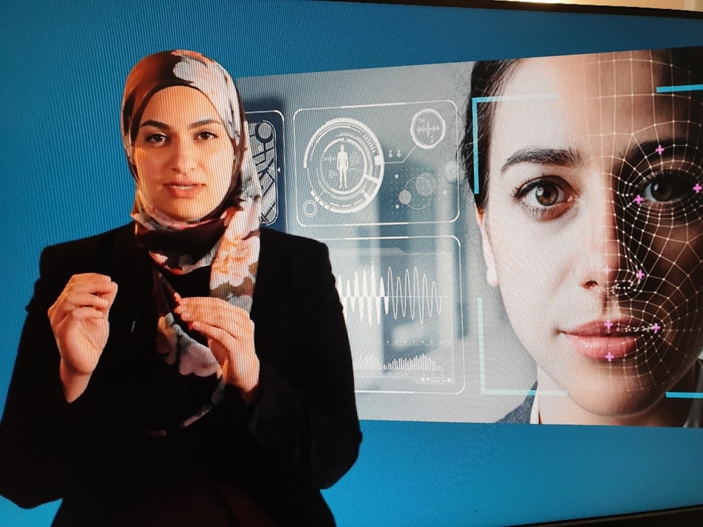 Female university student wearing headscarf standing in front of screen showing a female face being scanned by Artificial Intelligence