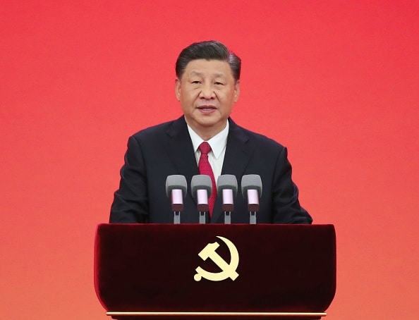 Chinese President Xi Jinping standing in front of a red backdrop at a podium with four identical microphones