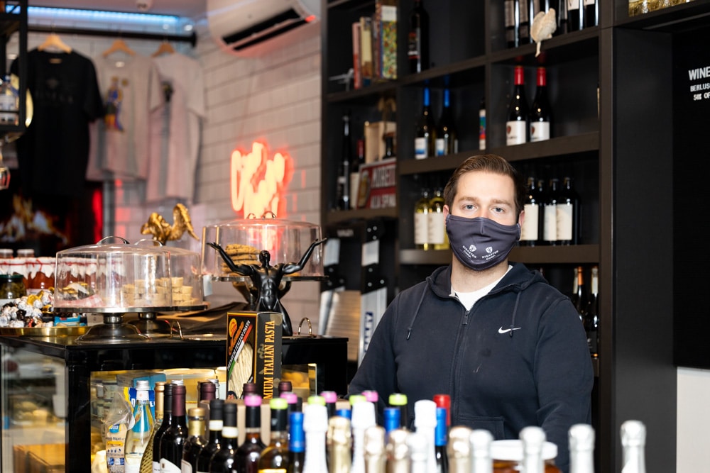 young male business owner wearing face mask standing behind counter of his deli cafe