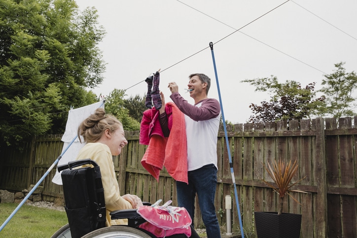 Mature man is hanging clothes on the washing line in his garden with his happy daughter sitting in her wheelchair next to him.