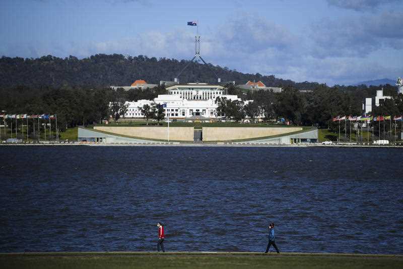 2 people are seen walking beside Lake Burley Griffin in Canberra with Parliament House in the background
