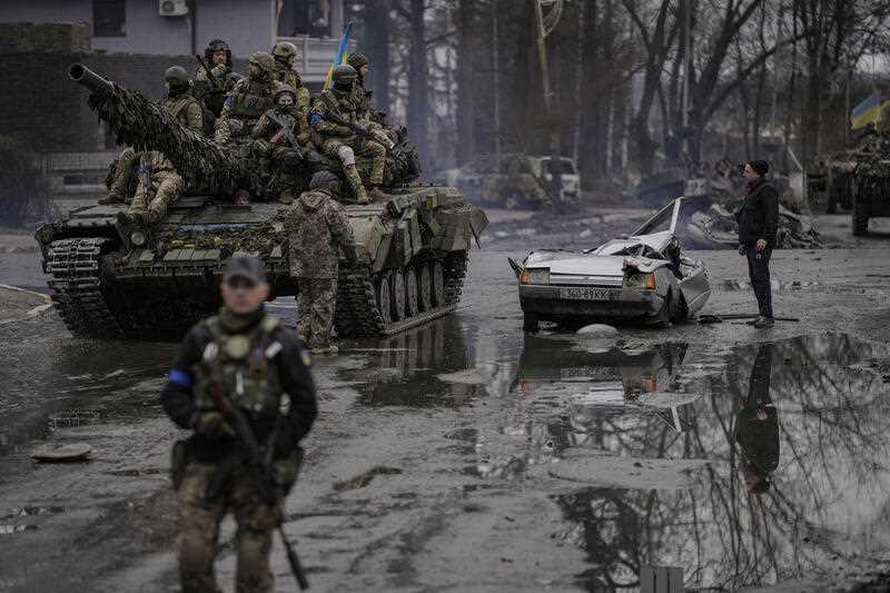 A man stands next to a civilian vehicle that was destroyed during fighting between Ukrainian and Russian forces that still contains the dead body of the driver as Ukrainian servicemen ride on a tank vehicle, outside Kyiv, Ukraine