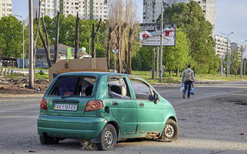 A man carries things as he walks on a street, past a damaged car, on the outskirts of Kharkiv, Ukraine, 14 May 2022