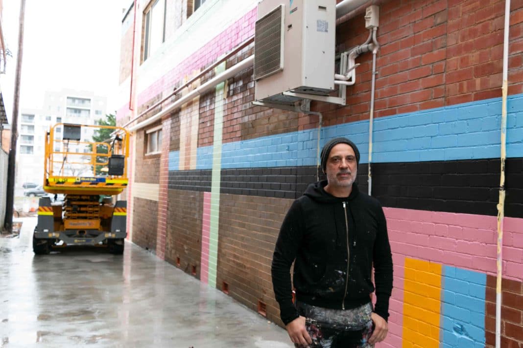 male artist in beanie standing beside mural he is painting in a laneway in Queanbeyan