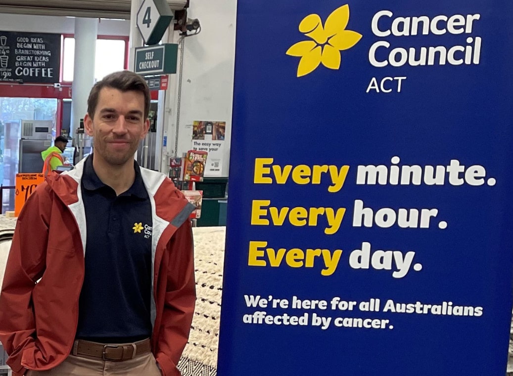 Smiling man in his 30s standing beside Cancer Council ACT signage in shopping centre