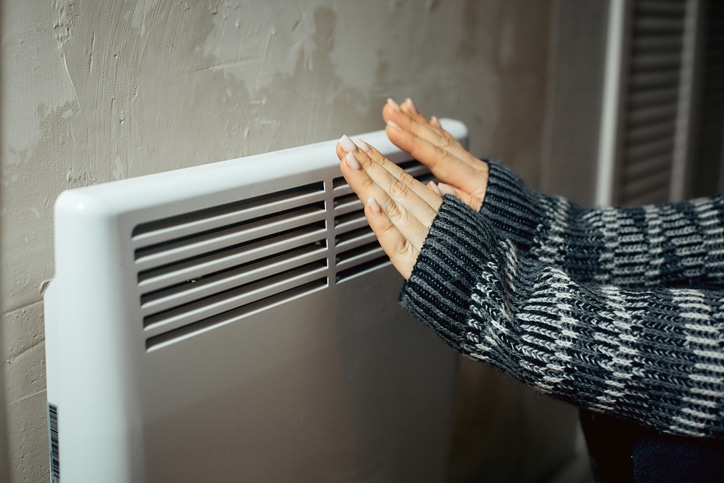 a woman warms her hands at the radiator in a cold house, problems with heating, heating the room with an electric convector