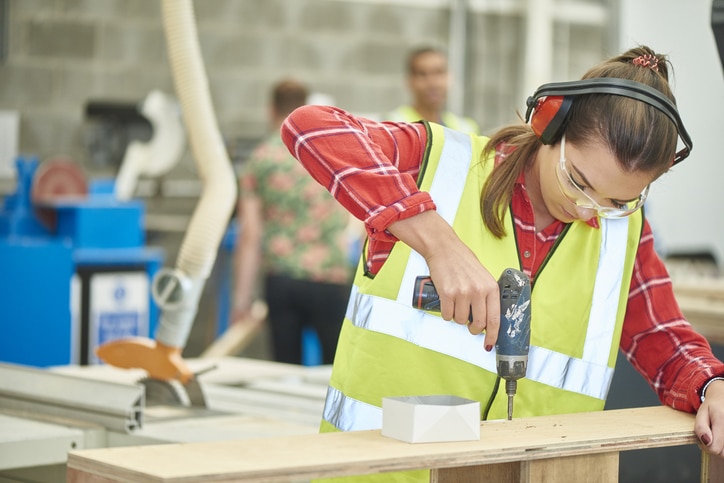 A young female carpenter looks to camera from her drill in a factory workshop. She is wearing ear defenders and safety goggles and drilling into a piece of timber