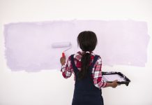 Back view of a woman painting the wall in purple, using a roller and a plastic tray for painting. The woman wearing a plaid shirt with overall jeans.